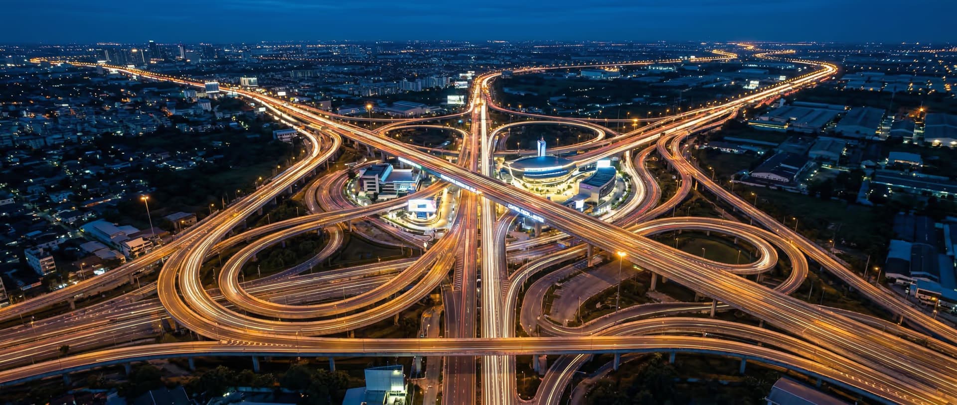 Bird's-eye aerial view of a multi-lane highway interchange at blue hour with light trails converging at a single point, representing GDV Holdings' centralized risk governance across multiple operating vectors